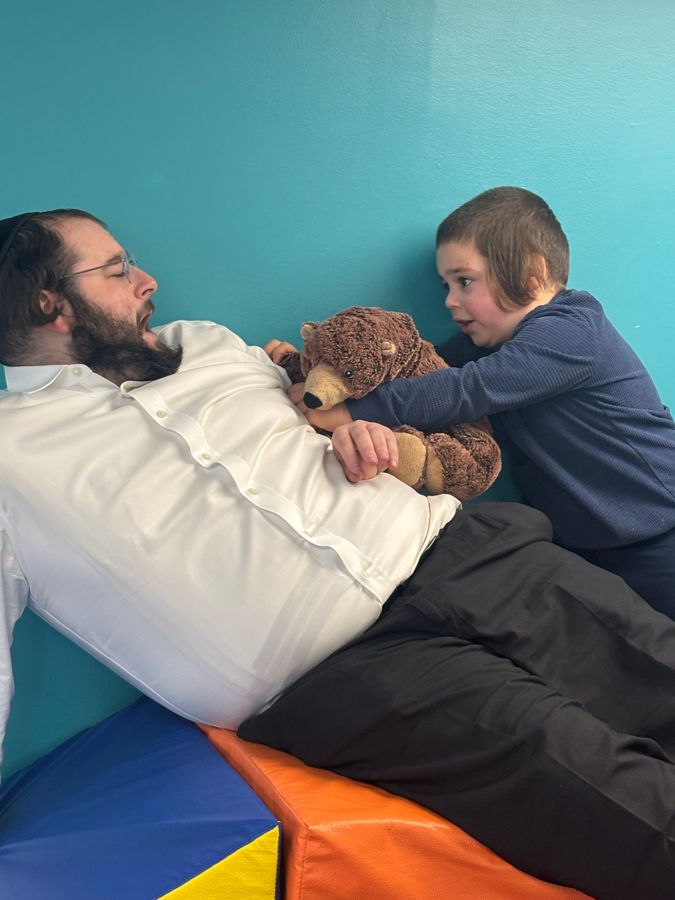 A boy tucking a teddy bear against a clinician pretending to sleep